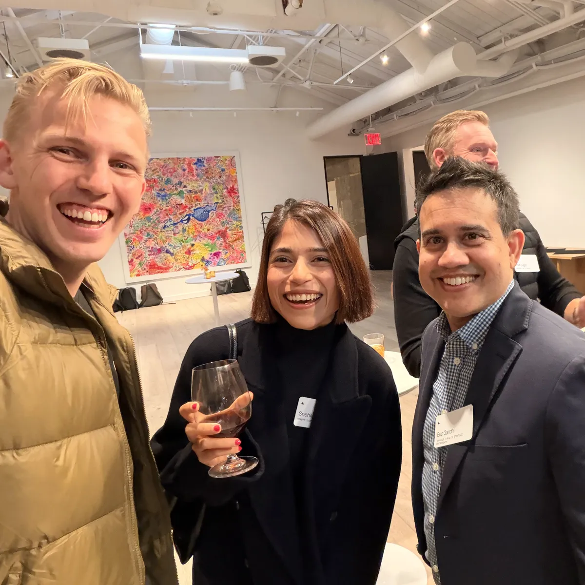 Selfie of Jono, Sne holding a glass of wine, and Eric Gandhi in a navy blazer, in front of a large colorful painting