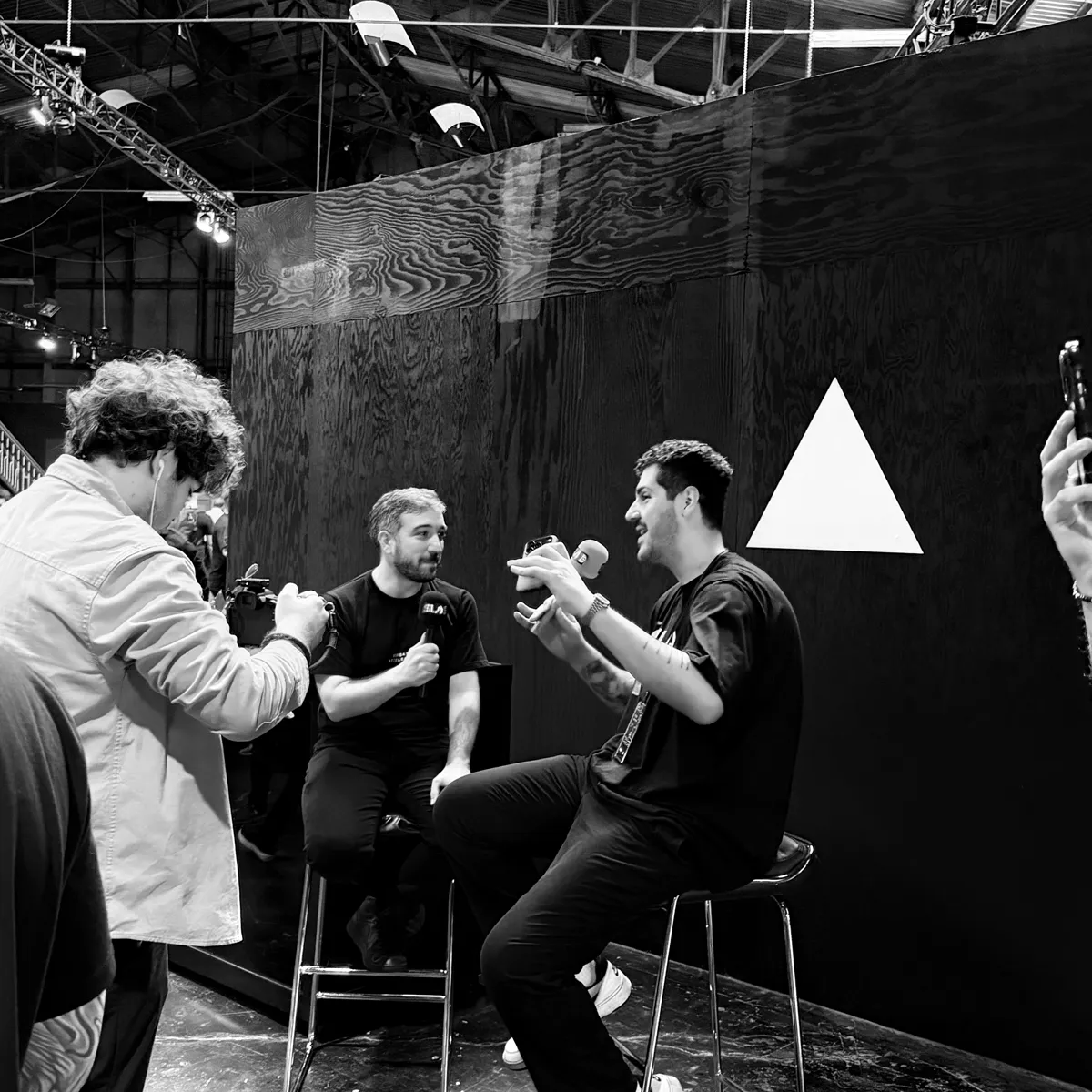 Black and white photo of a live interview with Evil Rabbit from Vercel on bar stools in front of a black wall with a white Vercel triangle, while a camera operator films