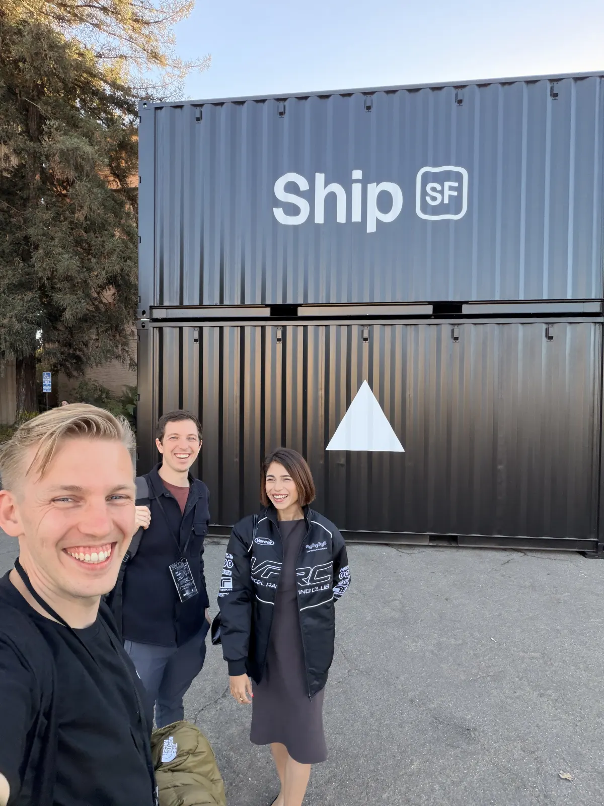 Selfie of Jono, Hal from Candid Leap, and Sne in a Vercel Racing Club jacket in front of two stacked black shipping containers printed with the Ship SF logo and the Vercel triangle