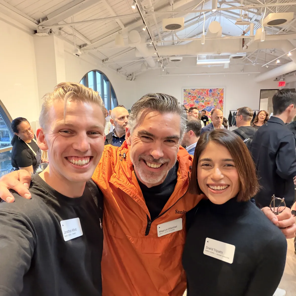 Selfie of Jono and Sne from Roboto Studio either side of Magnus Hillestad, who is wearing an orange Sanity jacket, in a busy room with a white beamed ceiling
