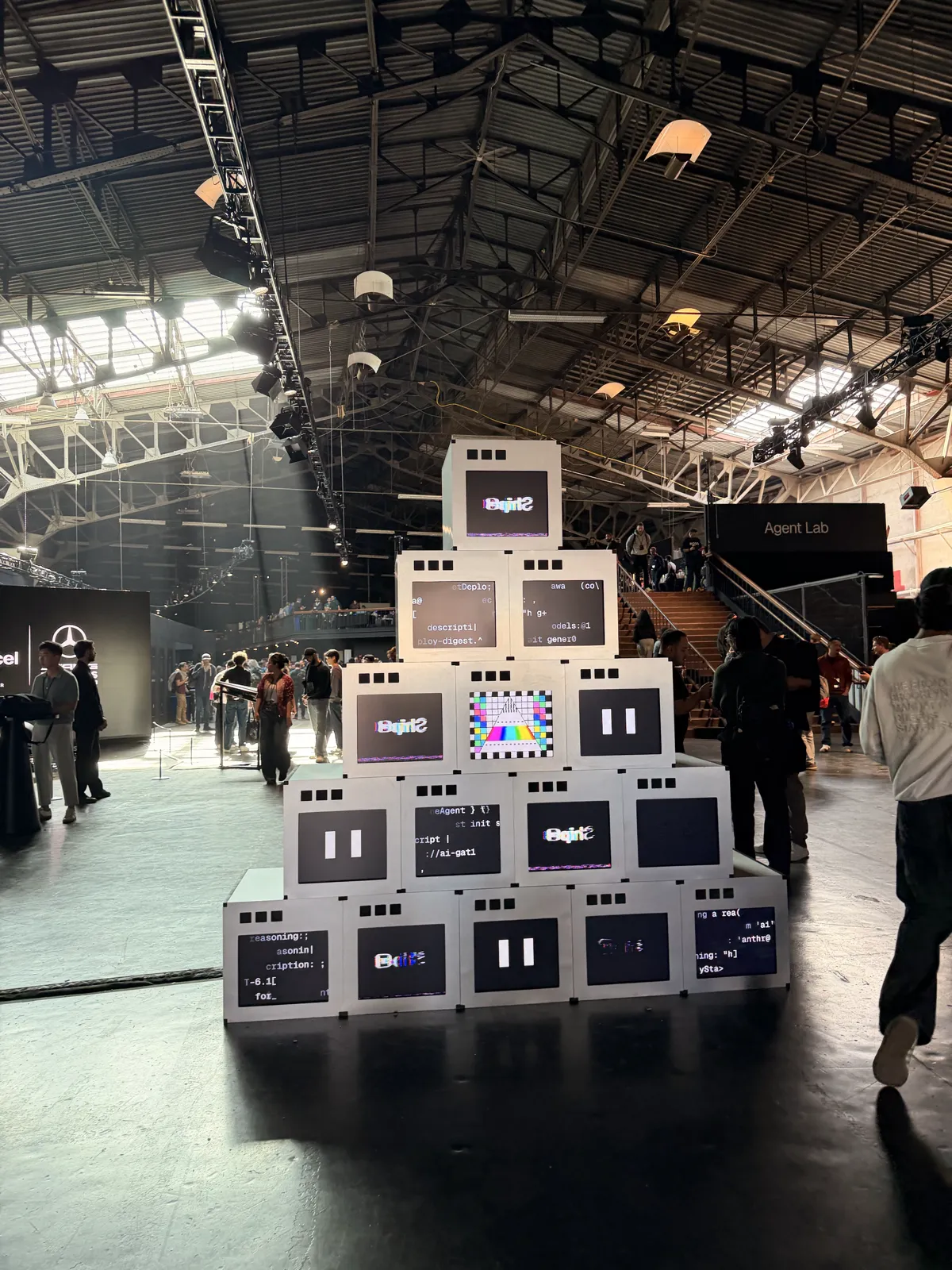 A pyramid of white boxy monitors showing code and Ship logos on the hangar floor at Vercel Ship, with the Agent Lab stairs behind it