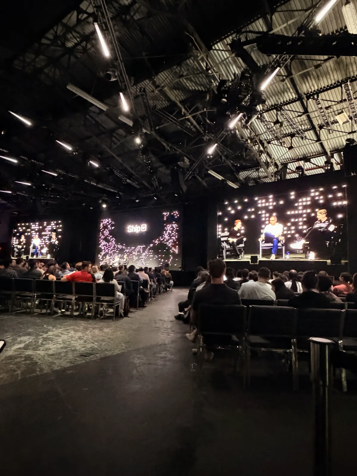 A packed audience watching a three-person panel on the main stage at Vercel Ship, under a hangar roof with strip lights and a Ship SF light wall