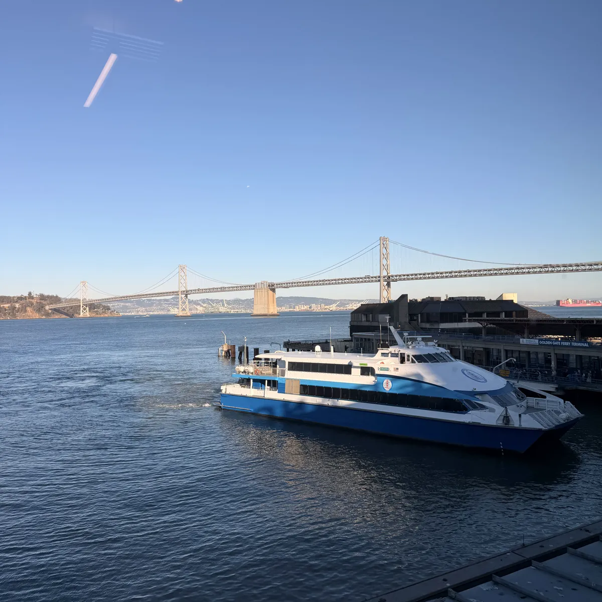 A blue and white ferry docked at the Golden Gate Ferry Terminal with the Bay Bridge behind it, seen from the partner event venue